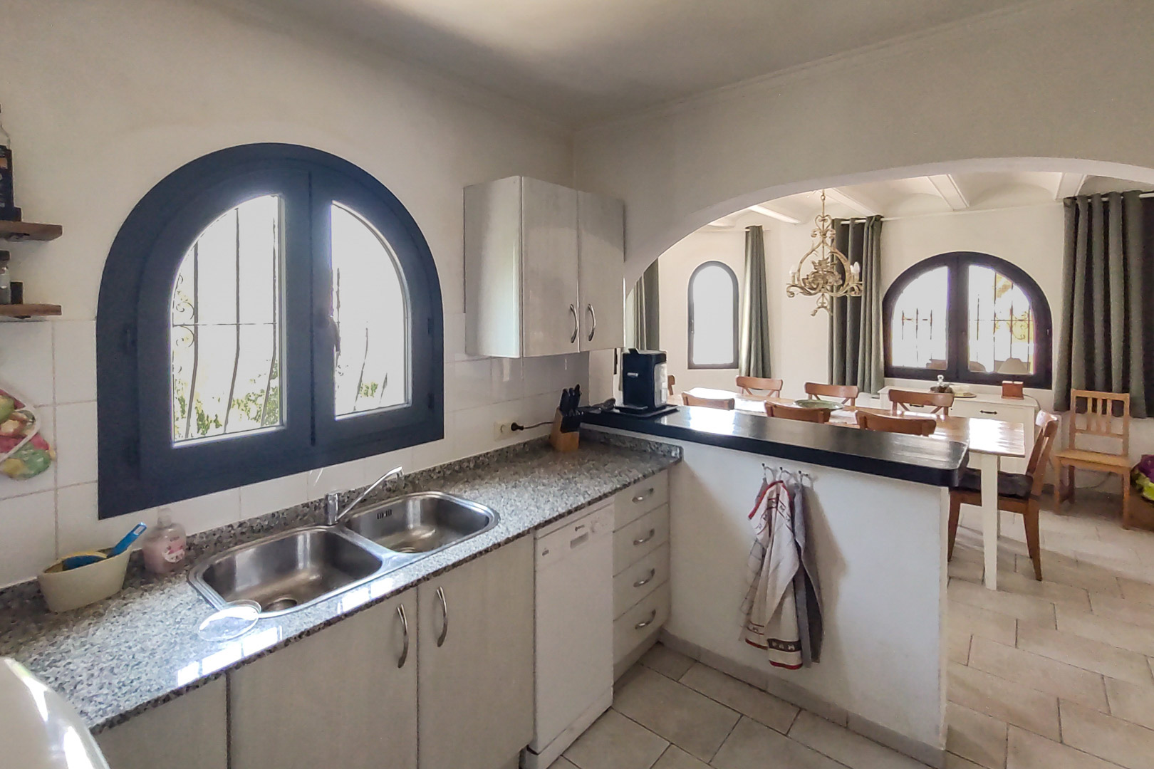 Cocina equipada con encimera de granito y fregadero doble, abierta al comedor mediante un arco. Incluye lavavajillas y una ventana de medio punto con carpintería oscura.