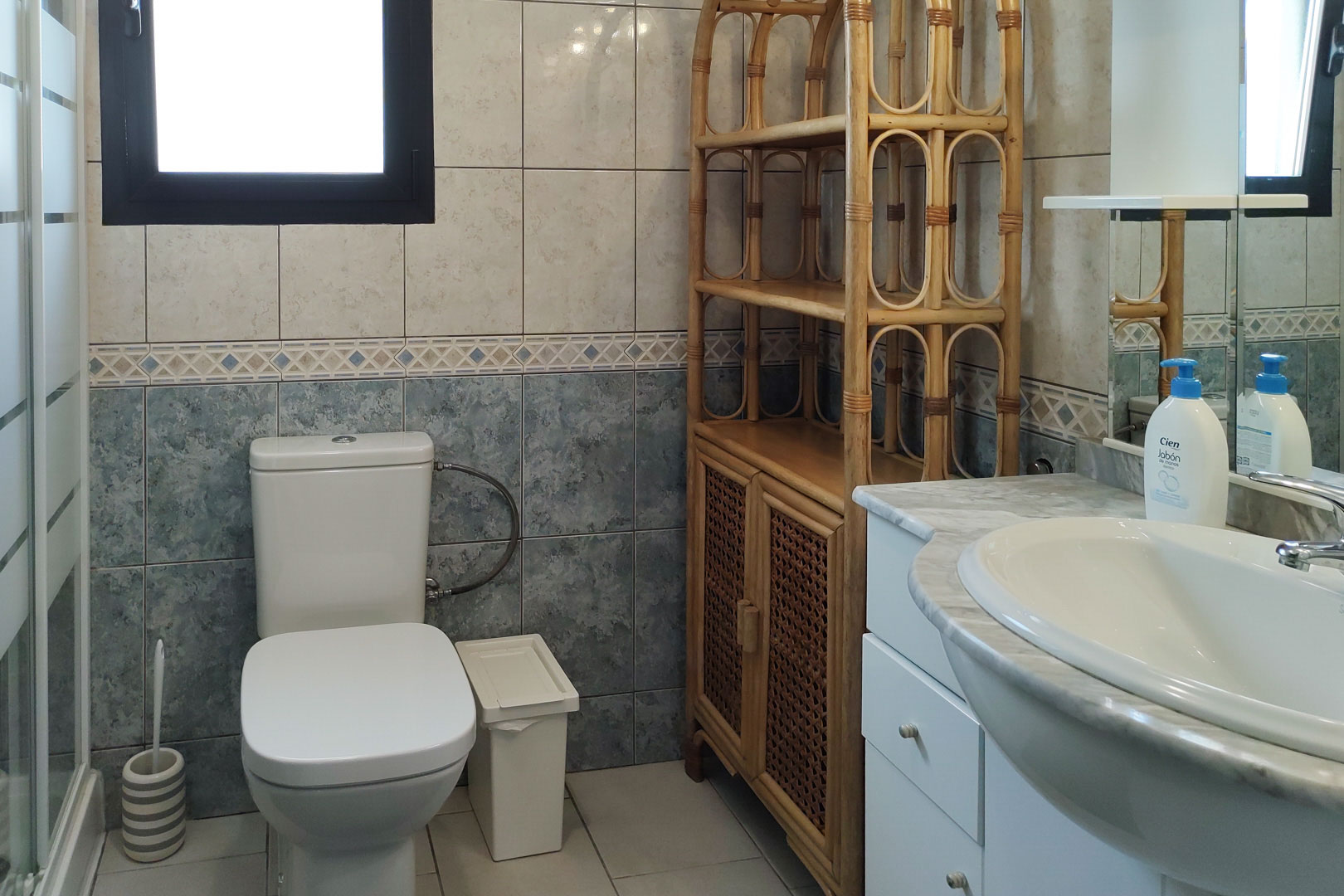 Cuarto de baño con encimera de mármol, estantería de bambú y paredes alicatadas en dos tonos con cenefa decorativa. Incluye luz natural y sanitarios blancos.