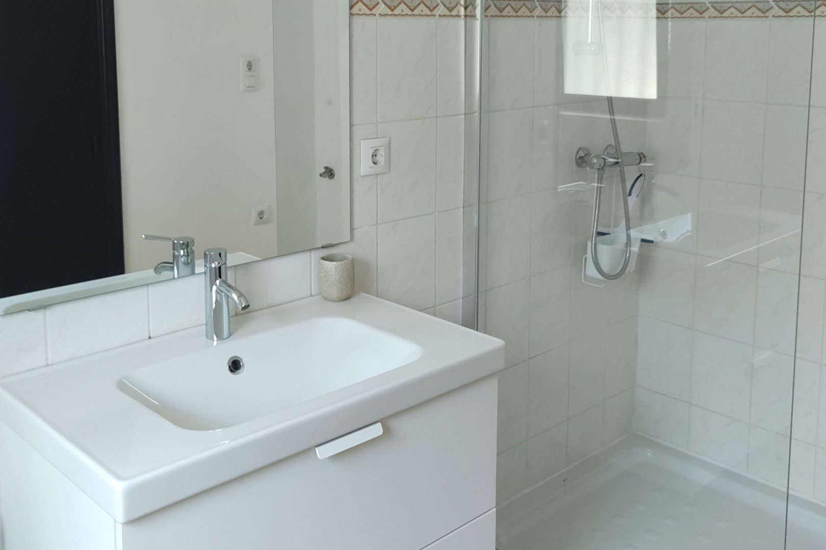 Cuarto de baño funcional con lavabo sobre mueble blanco, grifería cromada y ducha con mampara de cristal. Paredes alicatadas en blanco con cenefa decorativa superior.