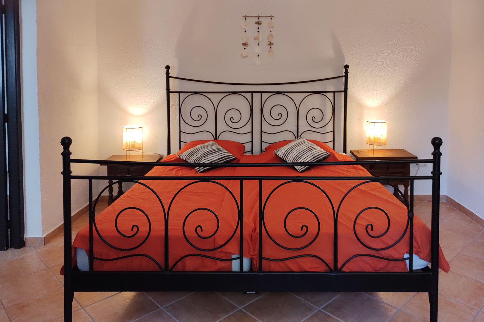 Dormitorio con cama de forja negra y espirales decorativas, mesitas de madera y suelos de baldosa cerámica. La iluminación cálida resalta las paredes blancas texturizadas.