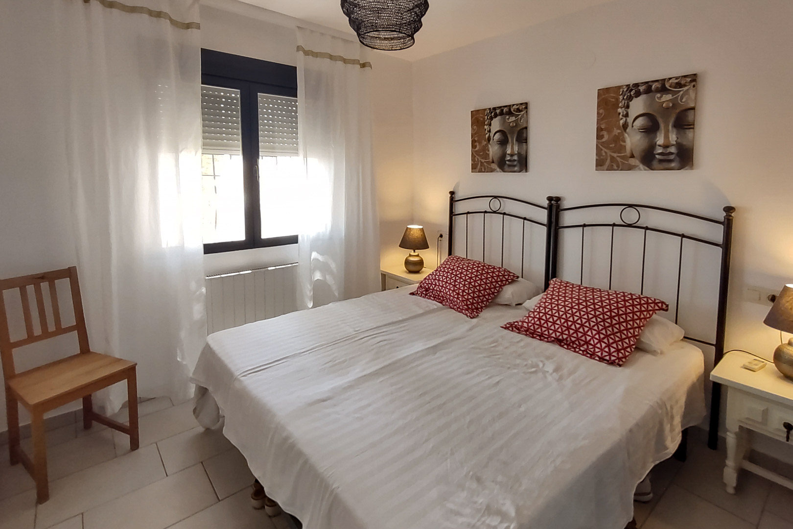 Dormitorio con camas individuales unidas, cabeceros de hierro forjado, cuadros decorativos de Buda y ventana con persianas y cortinas blancas.