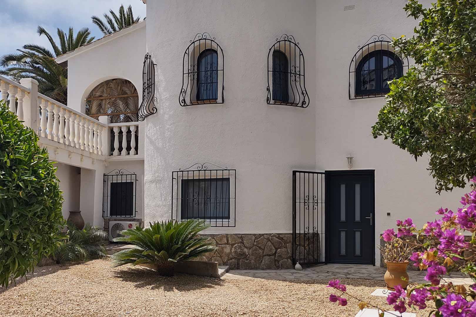 Fachada blanca de estilo mediterráneo con zócalo de piedra, ventanas protegidas por rejas de hierro forjado y entrada principal junto a un jardín de grava con buganvillas.