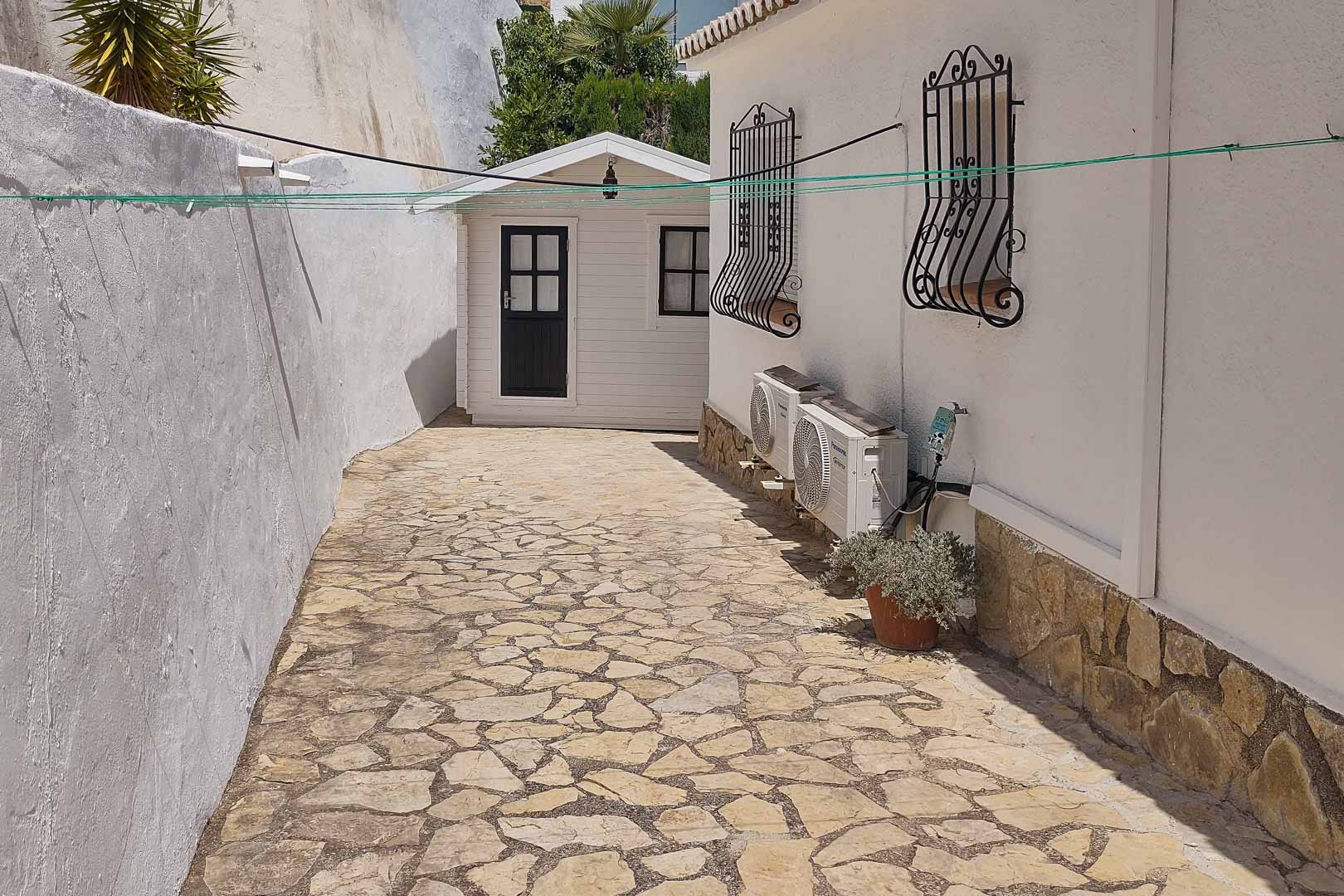 Pasillo lateral pavimentado en piedra natural que conduce a un trastero de madera independiente, junto a la fachada con equipos de climatización y rejas decorativas.