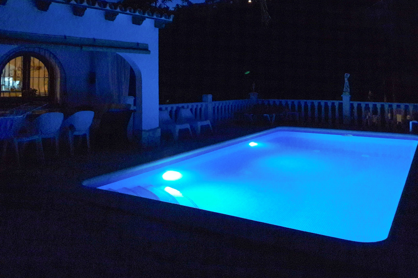 Piscina rectangular con iluminación nocturna LED en azul intenso, situada junto a un porche de arcos con zona de comedor exterior y balaustrada blanca.