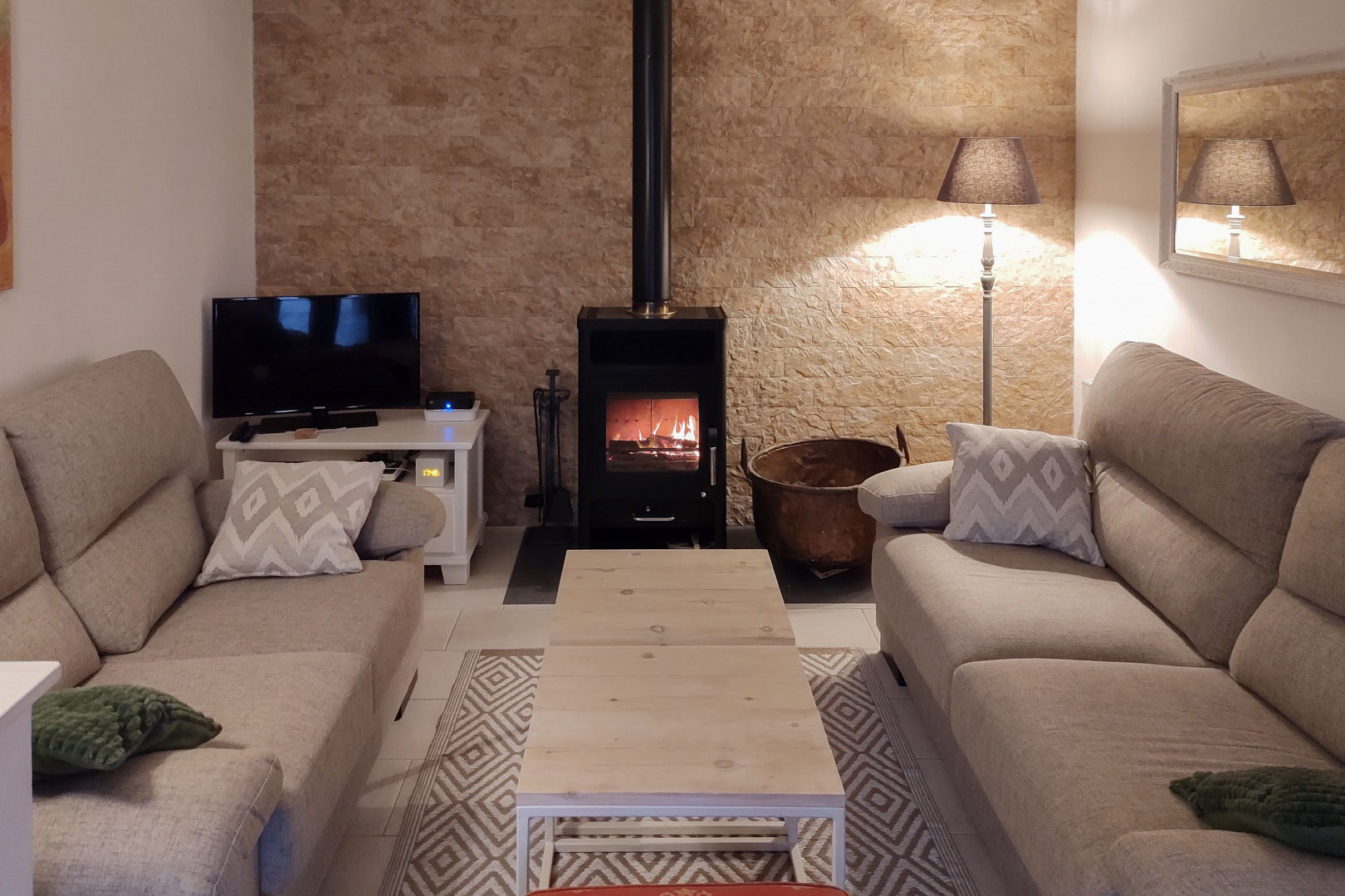 Salón acogedor con estufa de leña central sobre pared de piedra natural, equipado con sofás en tonos neutros y mesa de madera clara.