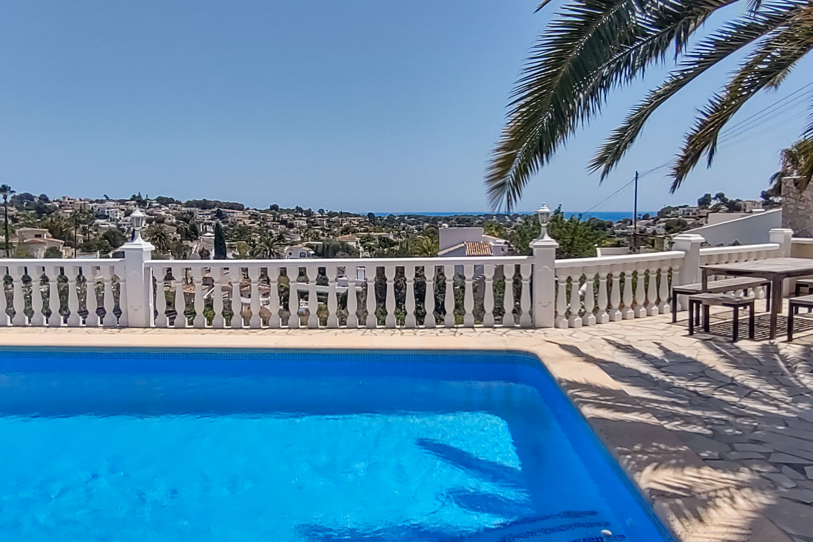 Terraza con piscina y balaustrada blanca que ofrece vistas panorámicas al mar y al valle, con zona de comedor exterior y pavimentación de piedra natural.