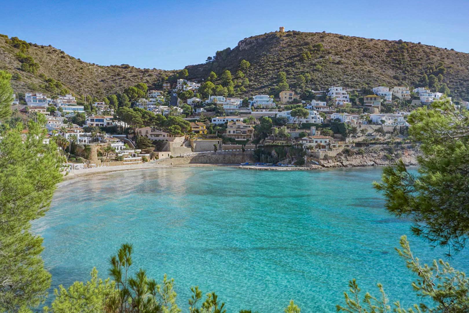 Vista panorámica de una cala mediterránea de aguas turquesas, rodeada de colinas con villas blancas y coronada por una torre de vigía histórica bajo un cielo despejado.