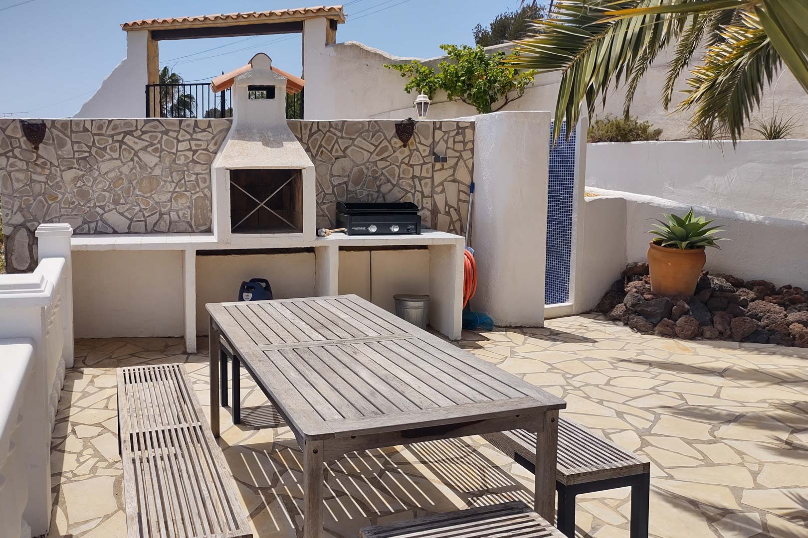 Zona de barbacoa de obra con plancha de gas, mesa de madera y bancos sobre suelo de piedra natural, junto a una ducha exterior revestida de gresite azul.
