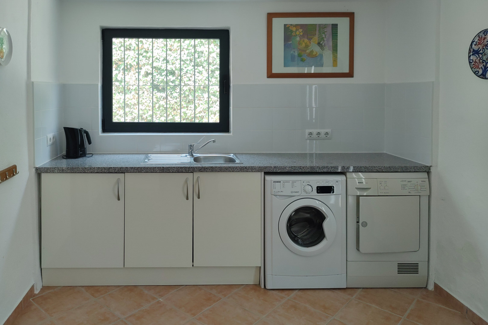 Zona de lavandería funcional con encimera de granito, fregadero de acero inoxidable, lavadora y secadora integradas bajo armarios blancos y suelo cerámico.