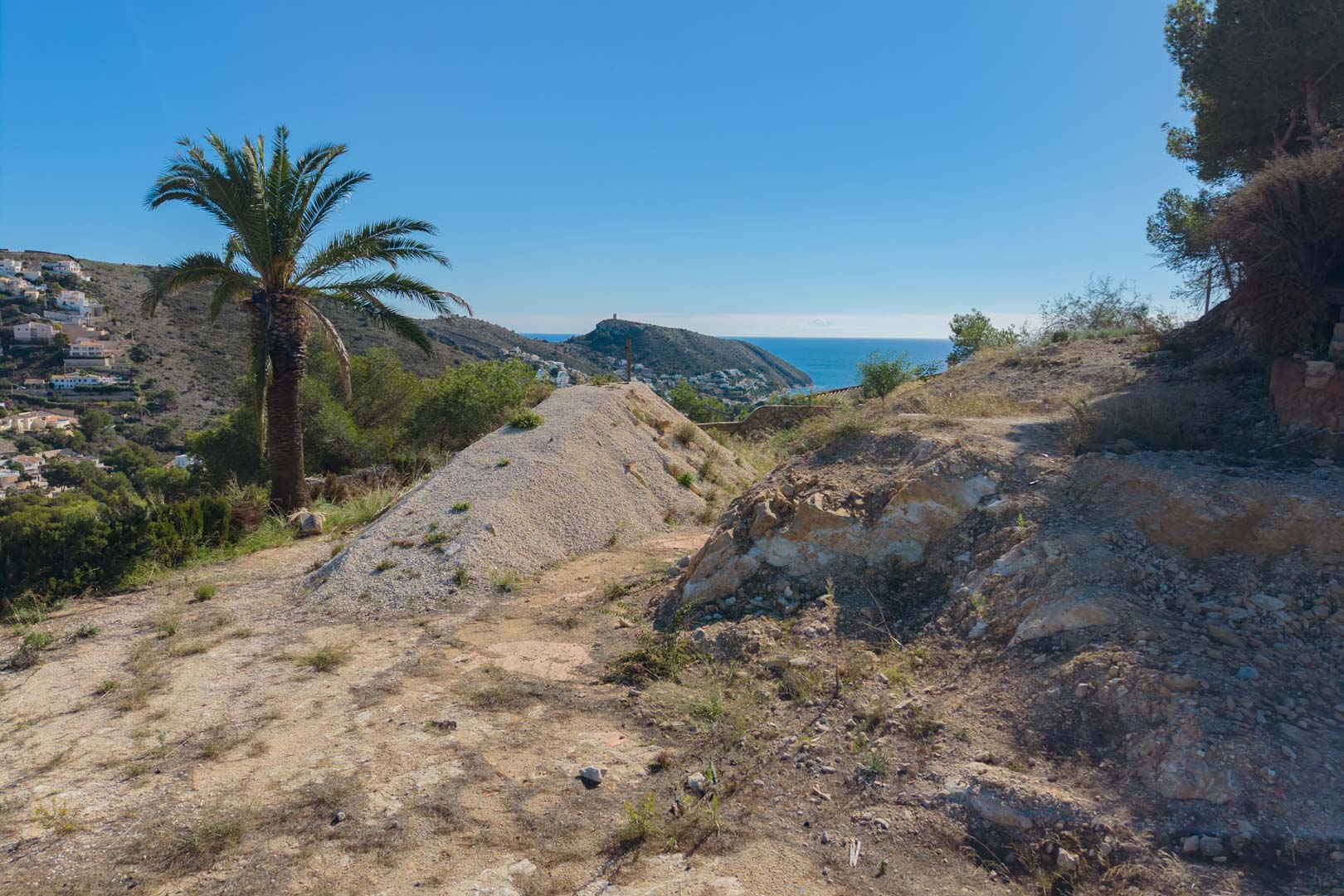 Parcela elevada en fase de preparación con vistas panorámicas al mar Mediterráneo y la costa de Moraira, rodeada de vegetación autóctona y palmeras.