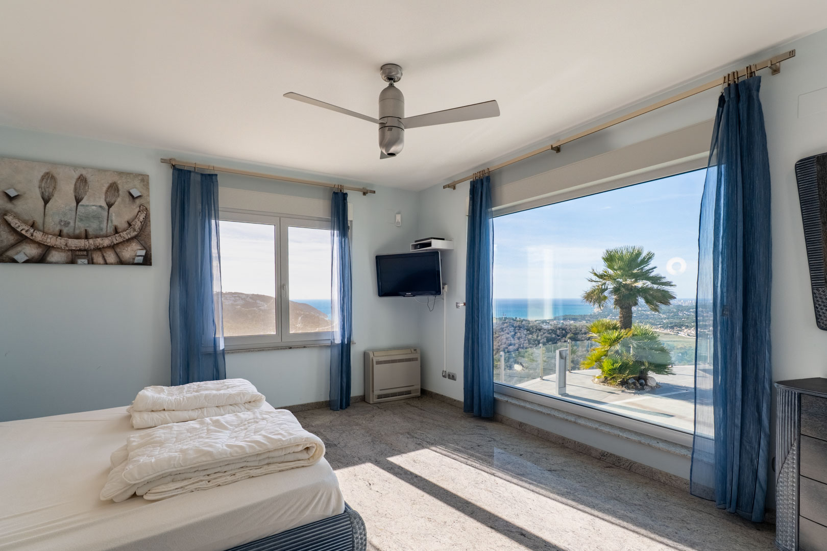Dormitorio luminoso con suelo de granito, cortinas azules y vistas al mar a través de amplios ventanales.
