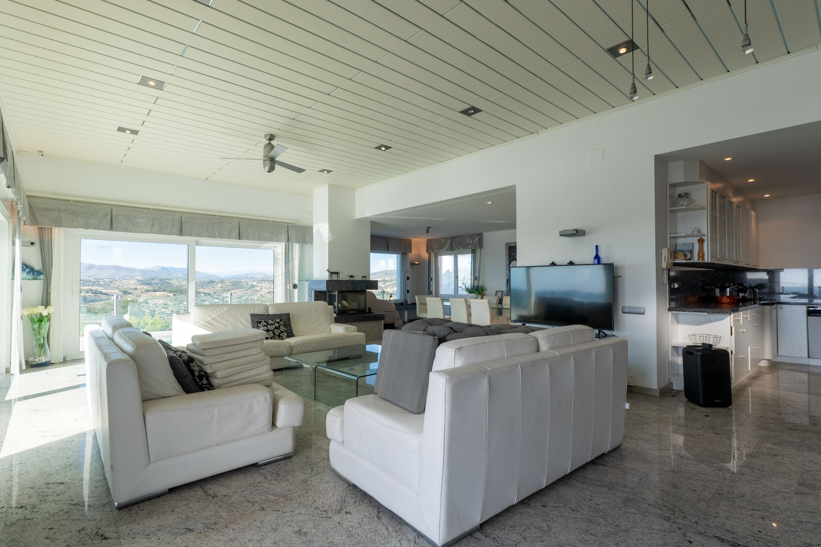 Luminoso salón con sofás de cuero blanco y vistas panorámicas. Suelos de granito pulido y acceso a la cocina.