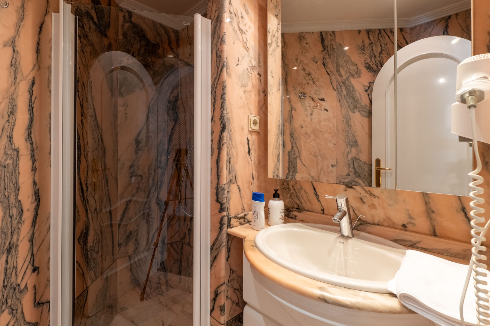 Baño revestido íntegramente en mármol rosa con vetas oscuras, equipado con ducha acristalada, lavabo encastrado sobre encimera de piedra y espejo de gran formato.
