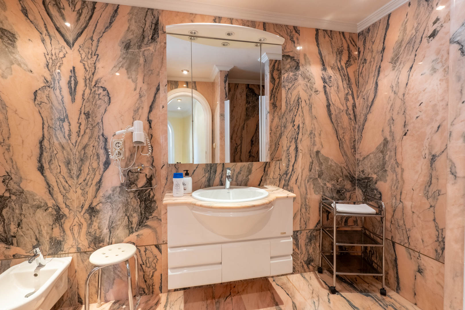 Baño revestido íntegramente en mármol rosa con vetas oscuras, equipado con mueble de lavabo blanco, espejo con iluminación integrada y bidé a juego.