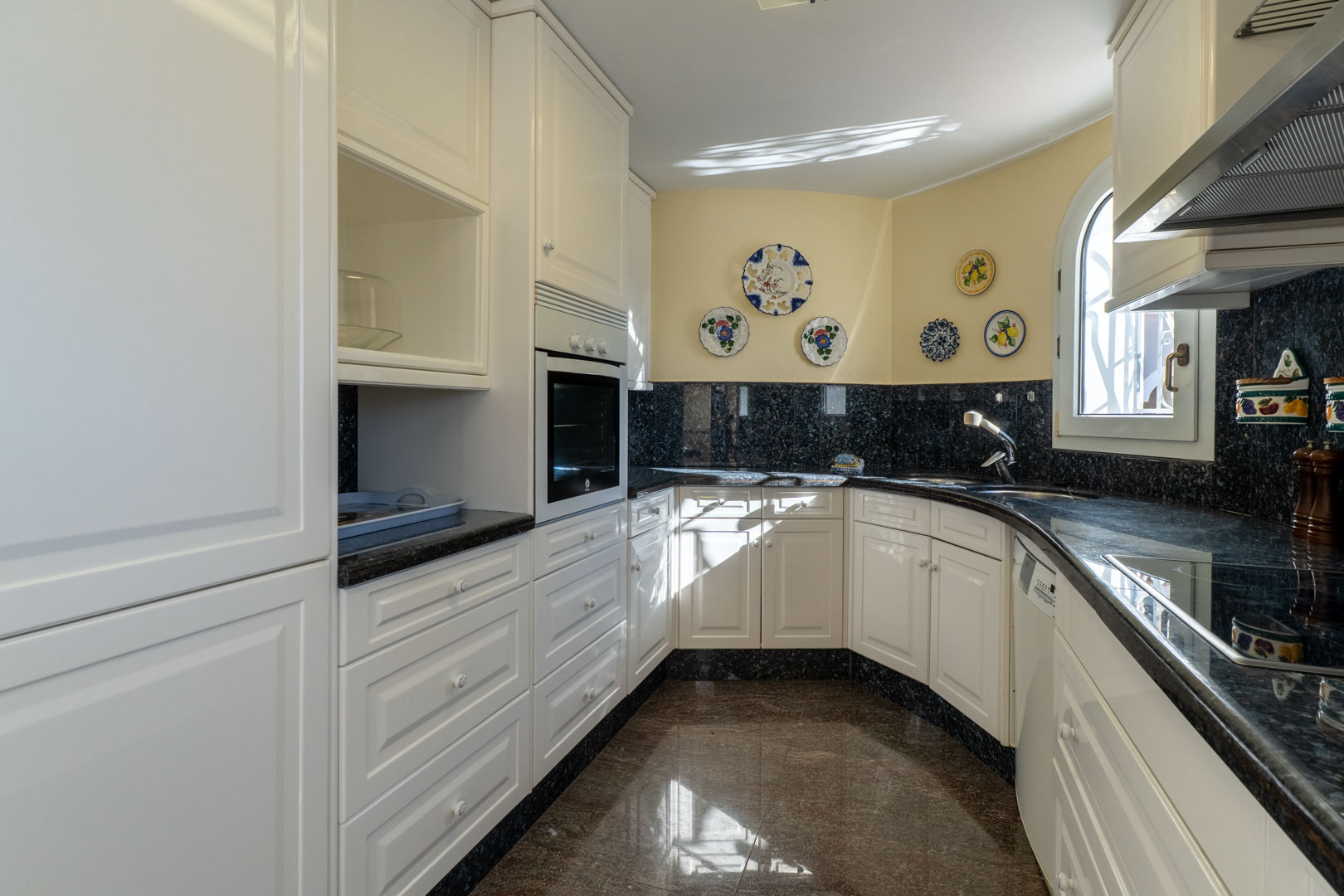 Cocina independiente con armarios blancos, encimera de granito oscuro y detalles cerámicos. Dispone de luz natural, ventana de arco y electrodomésticos integrados.