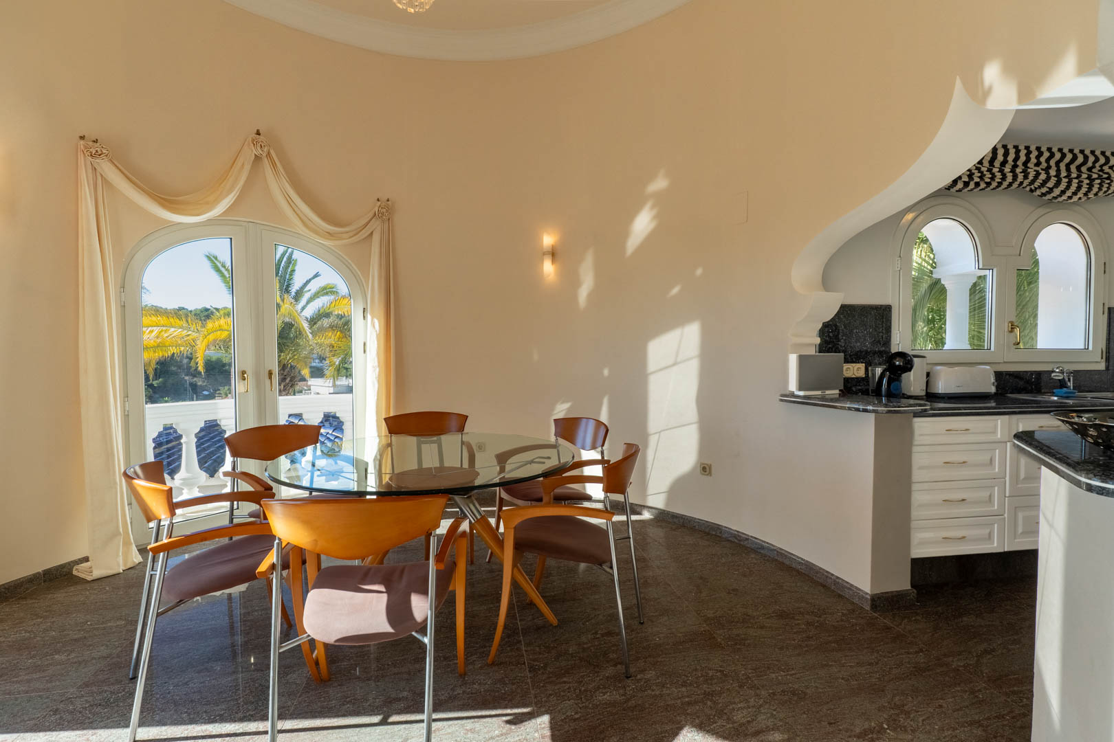 Comedor circular con mesa de cristal y sillas de madera, integrado junto a una cocina con encimera de granito y acceso directo a la terraza mediante puertas de arco.