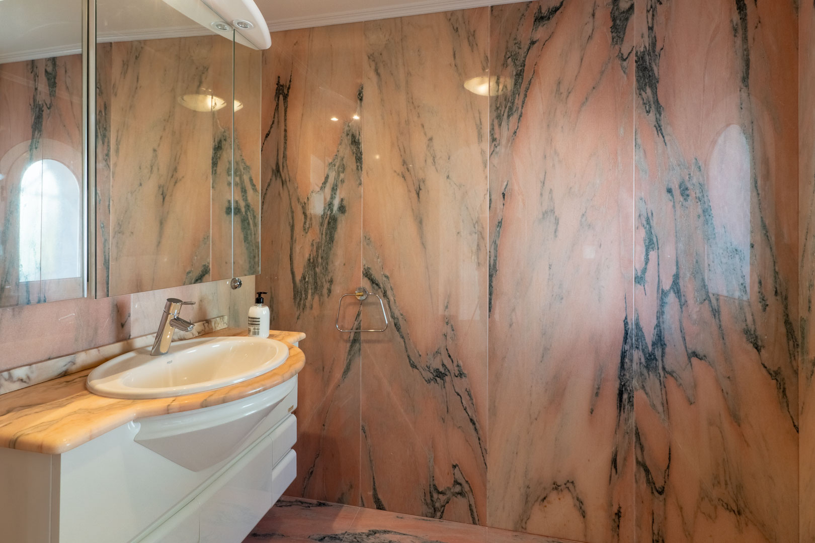 Cuarto de baño con revestimiento integral de mármol rosa y vetas oscuras. Incluye un lavabo encastrado en encimera de piedra, mueble blanco y un gran espejo frontal.