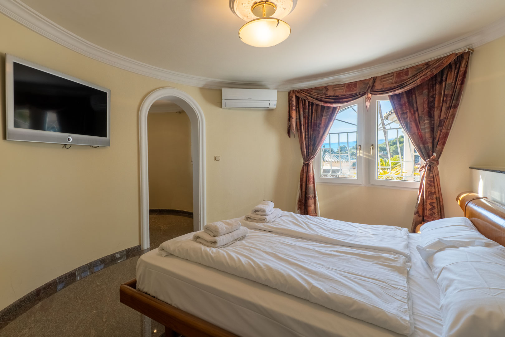 Dormitorio con paredes curvas, cama doble y acceso mediante arco. Incluye televisión de pared, aire acondicionado y ventana con vistas al mar y cortinajes clásicos.