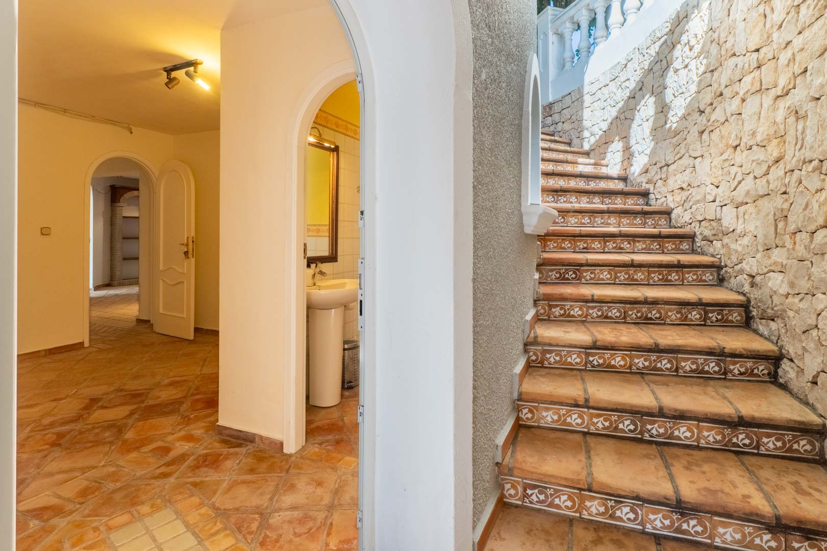 Escalera exterior con azulejos decorativos y muro de piedra natural, conectando con un pasillo interior de suelos de terracota y puertas en arco.