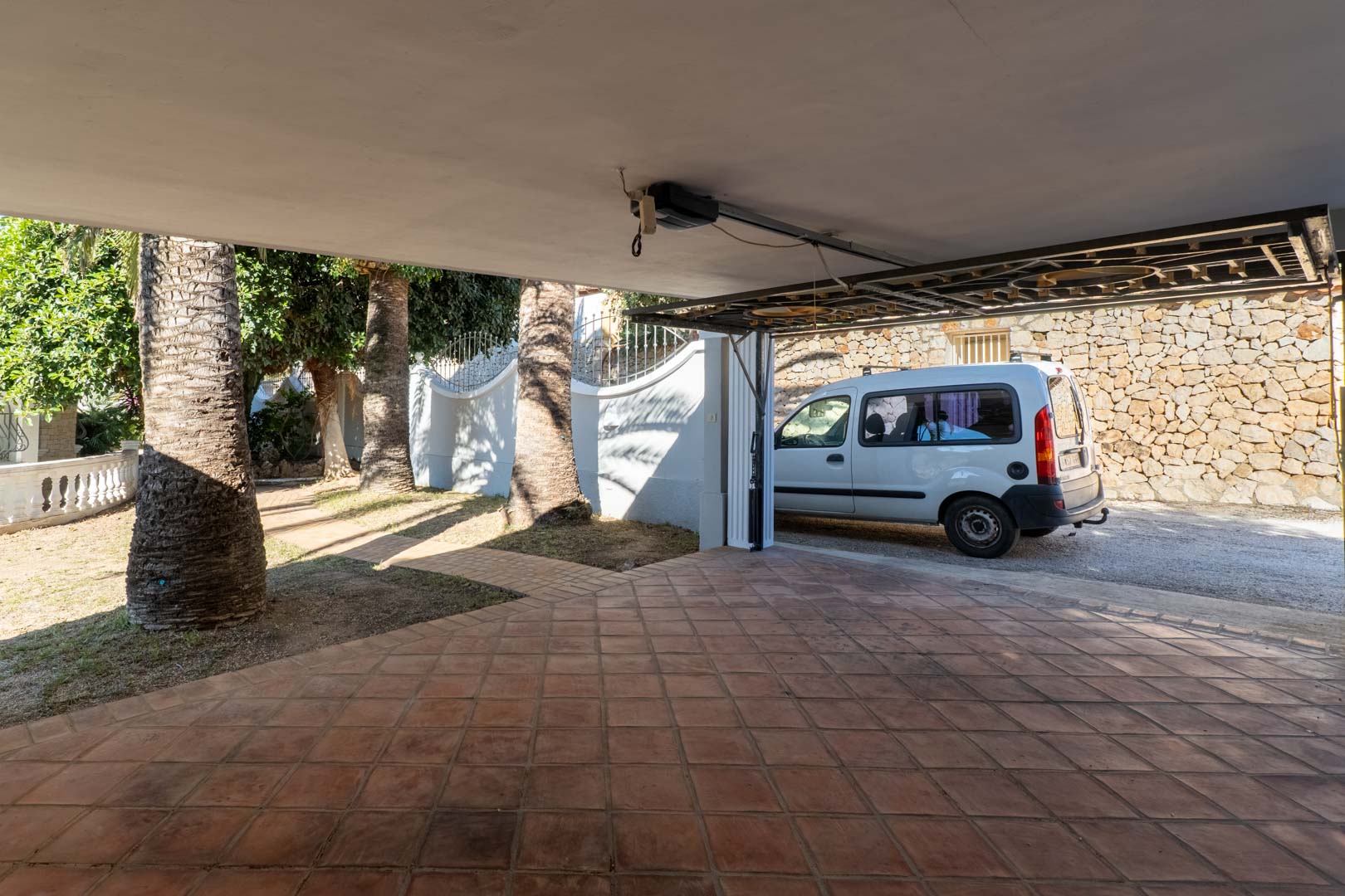 Garaje cubierto con suelo de baldosas de barro y puerta automática, con acceso directo a un jardín mediterráneo con palmeras y muros de piedra natural.