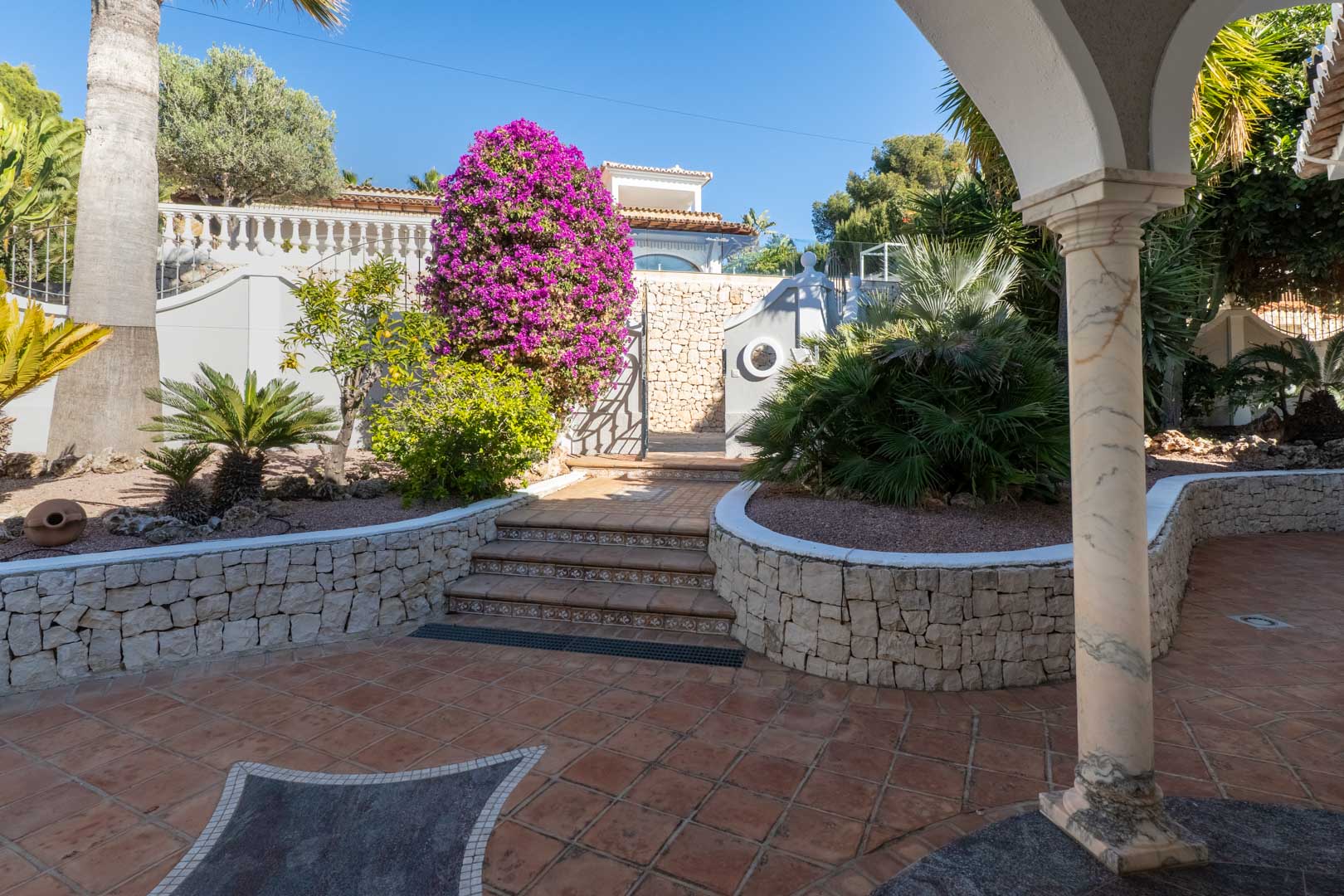 Patio exterior con suelos de terracota, muros de piedra seca y una buganvilla vibrante. Una columna de mármol enmarca el acceso a los cuidados jardines mediterráneos.