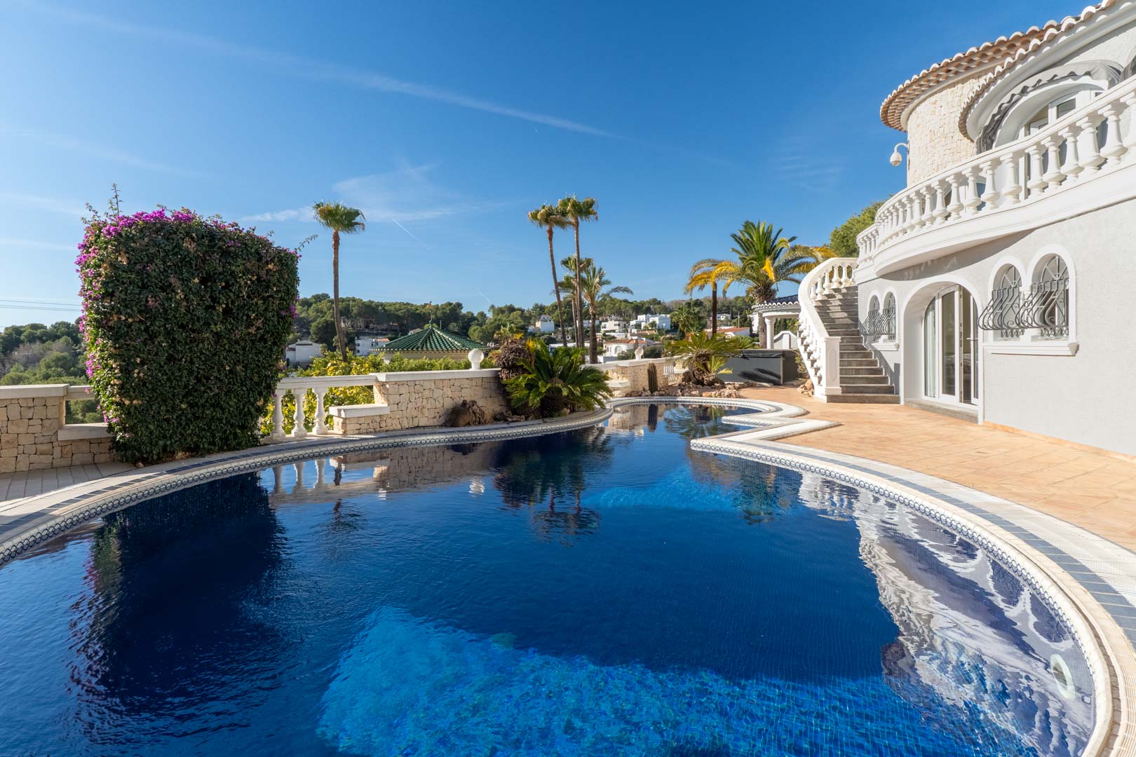Piscina de forma orgánica con gresite azul, rodeada de una terraza de gres y muros de piedra natural, junto a la fachada blanca de estilo mediterráneo con arcos y balaustradas.