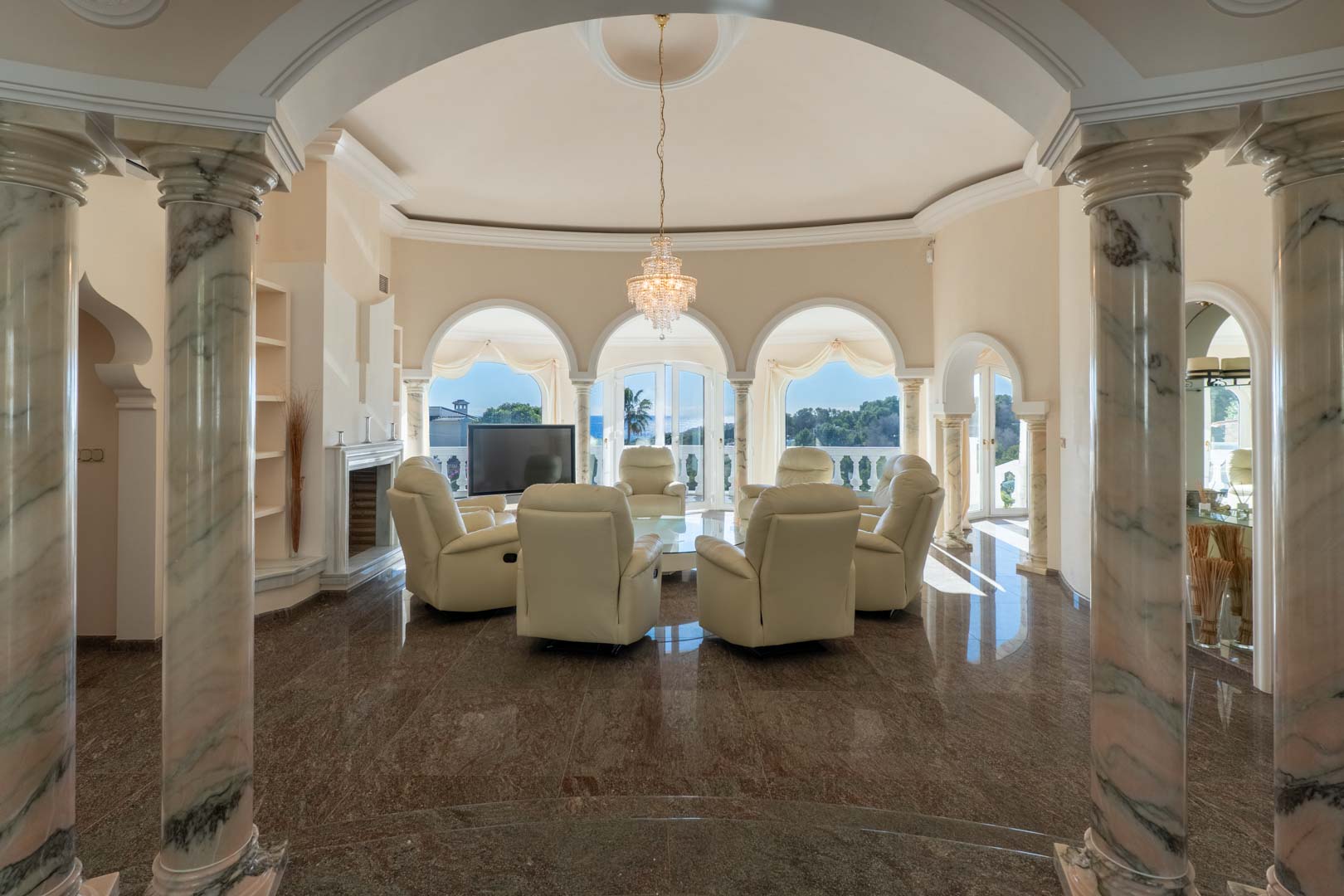 Salón circular con columnas de mármol veteado, suelos de granito pulido y ventanales en arco con vistas al mar Mediterráneo y zona de estar con sillones de piel blanca.