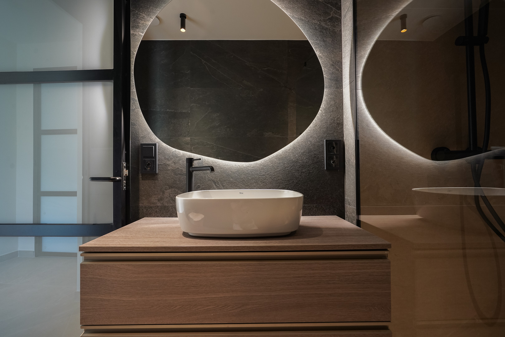Baño de diseño con lavabo sobre encimera, mueble de madera, espejo circular retroiluminado y paredes de gres oscuro con grifería en negro mate.