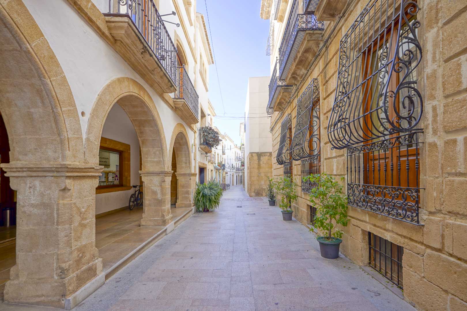 Calle peatonal en el centro histórico de Jávea con arcos de piedra tosca, balcones de forja y fachadas tradicionales mediterráneas.