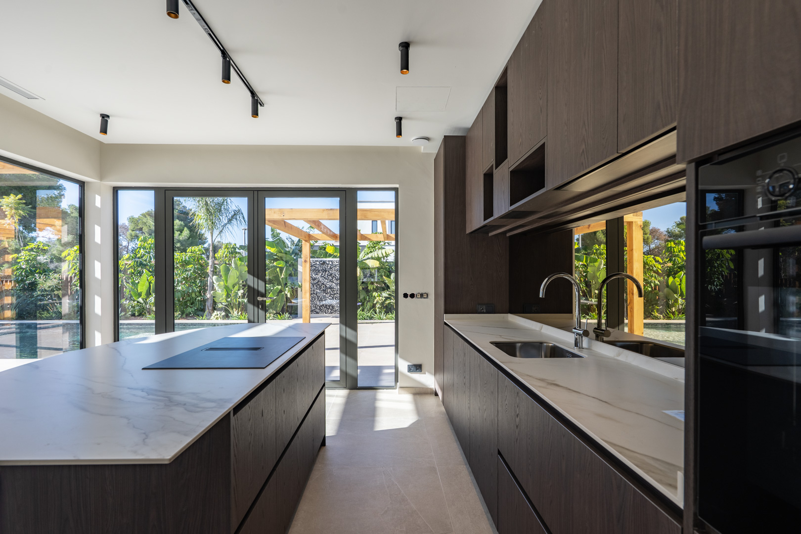 Cocina de diseño con isla central, encimeras de efecto mármol y armarios de madera oscura. Los ventanales ofrecen acceso directo al jardín y la zona de piscina.