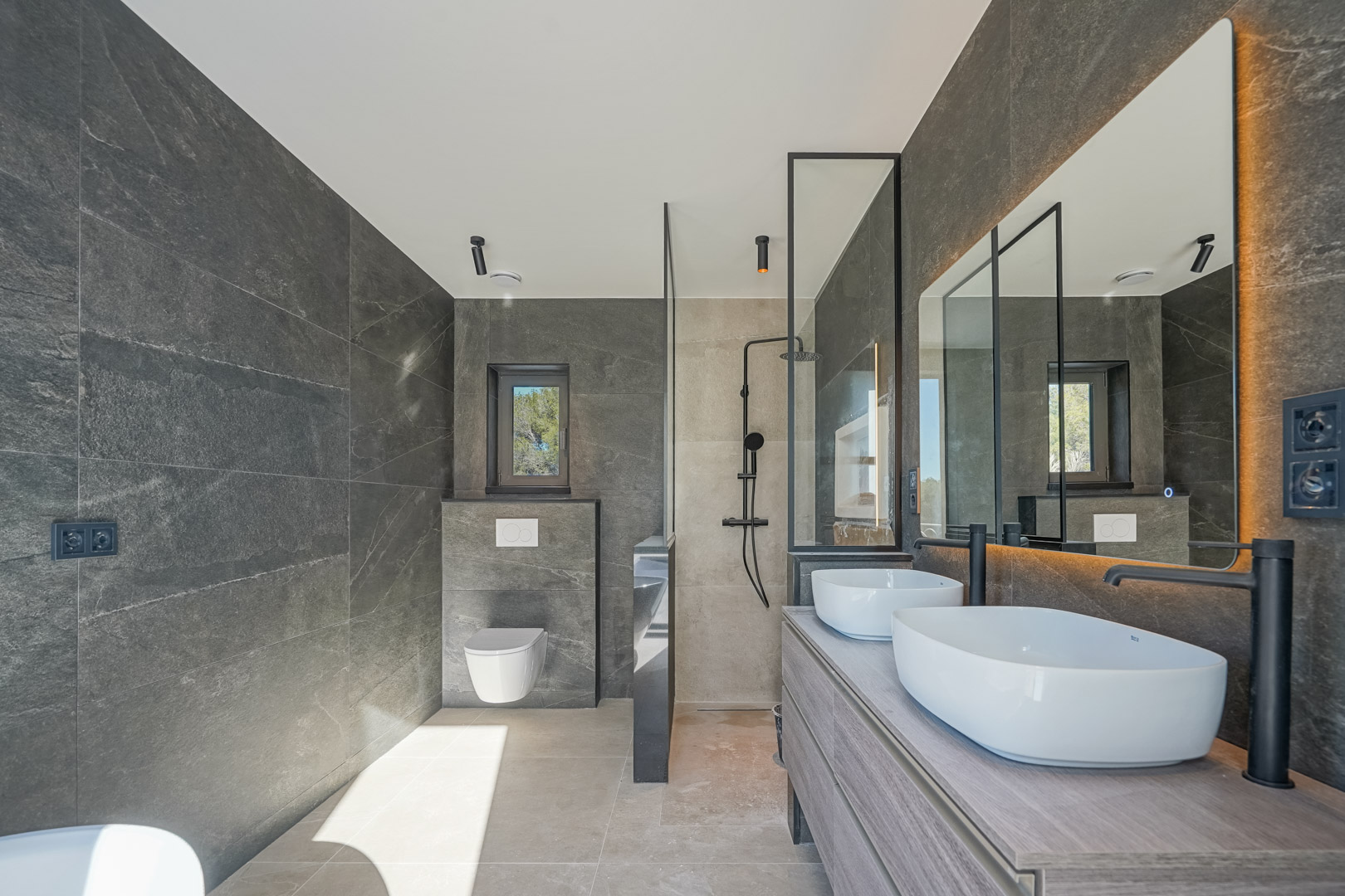 Cuarto de baño moderno con azulejos de piedra gris, lavabo doble sobre mueble de madera, grifería negra mate y ducha de obra con mampara de cristal.