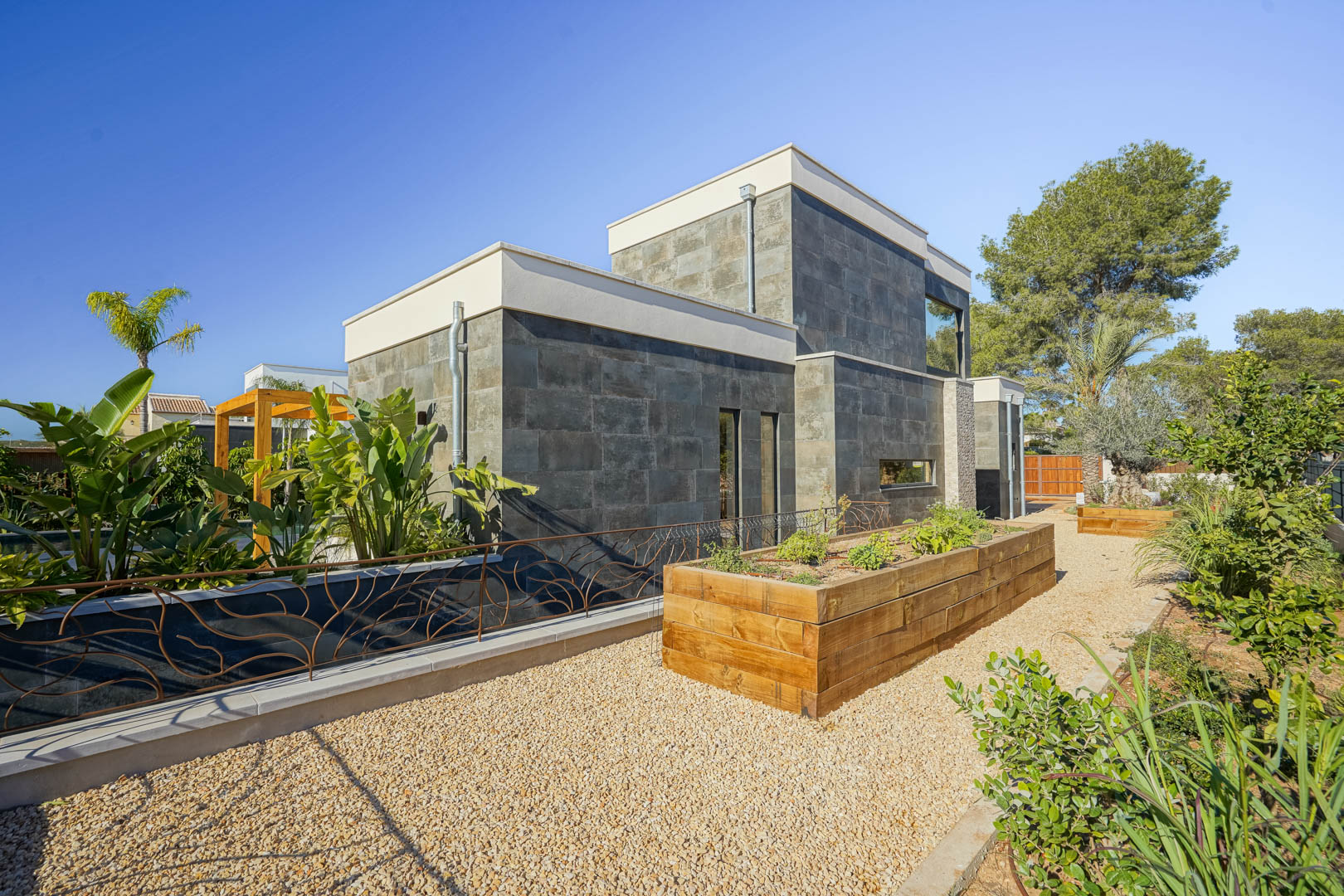 Fachada moderna revestida en piedra natural gris con jardineras de madera elevadas y senderos de grava en un entorno mediterráneo.