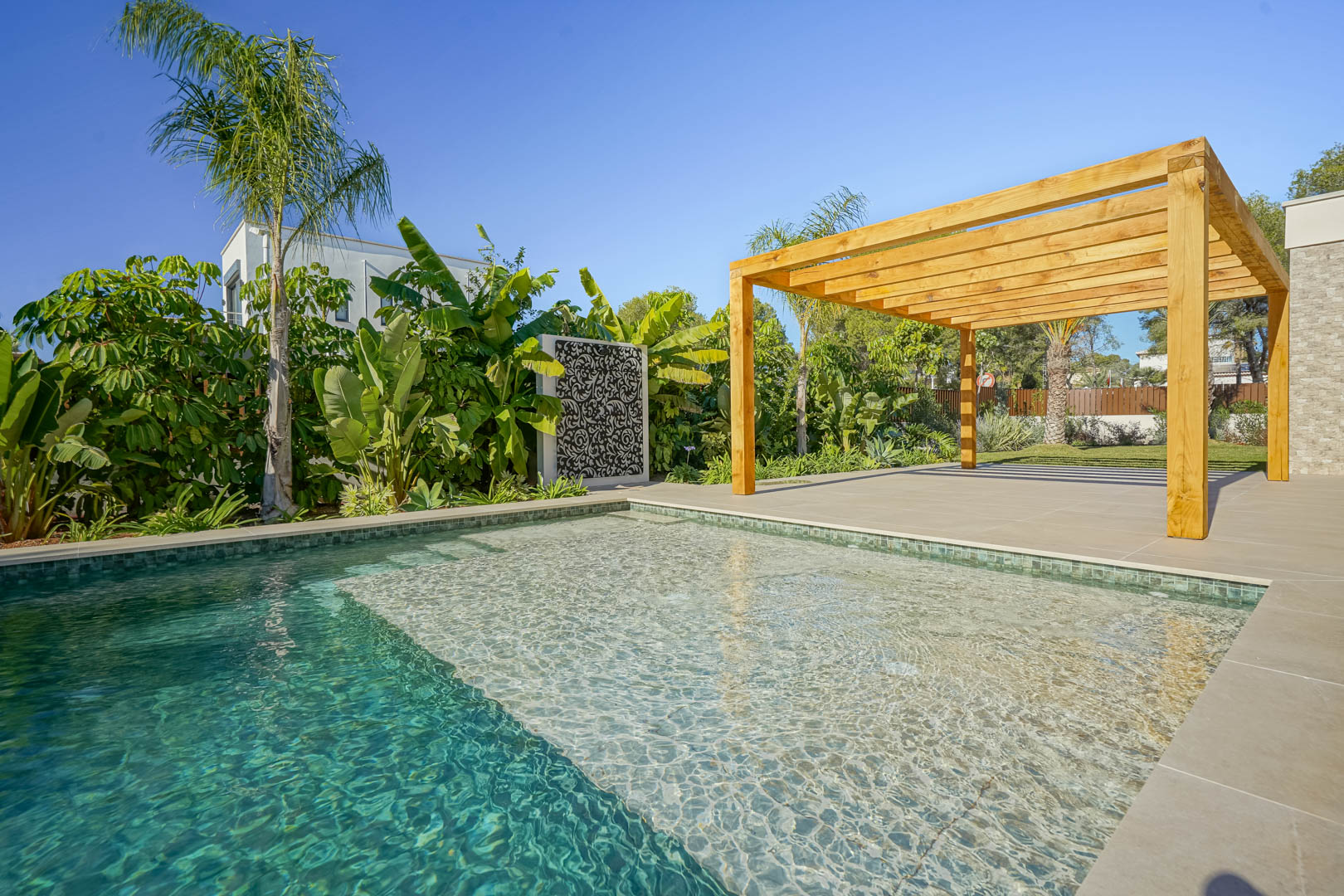 Piscina de estilo Bali con zona de playa, rodeada de una terraza de gres porcelánico y una pérgola de madera de iroko junto a un jardín mediterráneo.