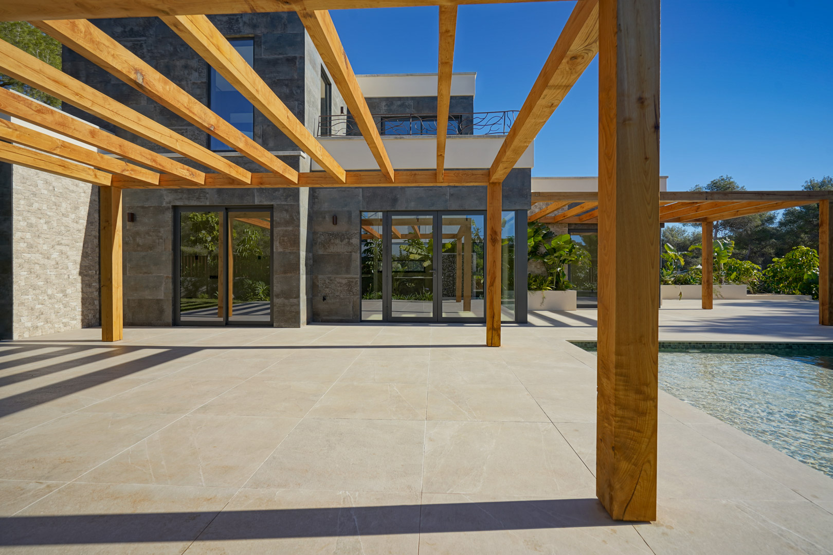 Terraza exterior con pérgola de madera de iroko sobre suelo porcelánico, junto a la piscina y fachada de piedra gris con amplios ventanales.