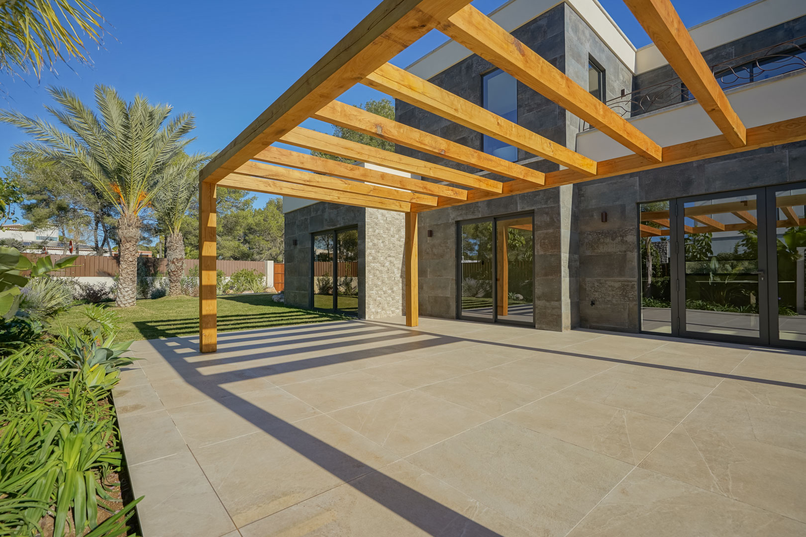 Terraza exterior con pérgola de madera de iroko y suelo de gres porcelánico, integrada en un jardín mediterráneo con fachada de piedra natural.
