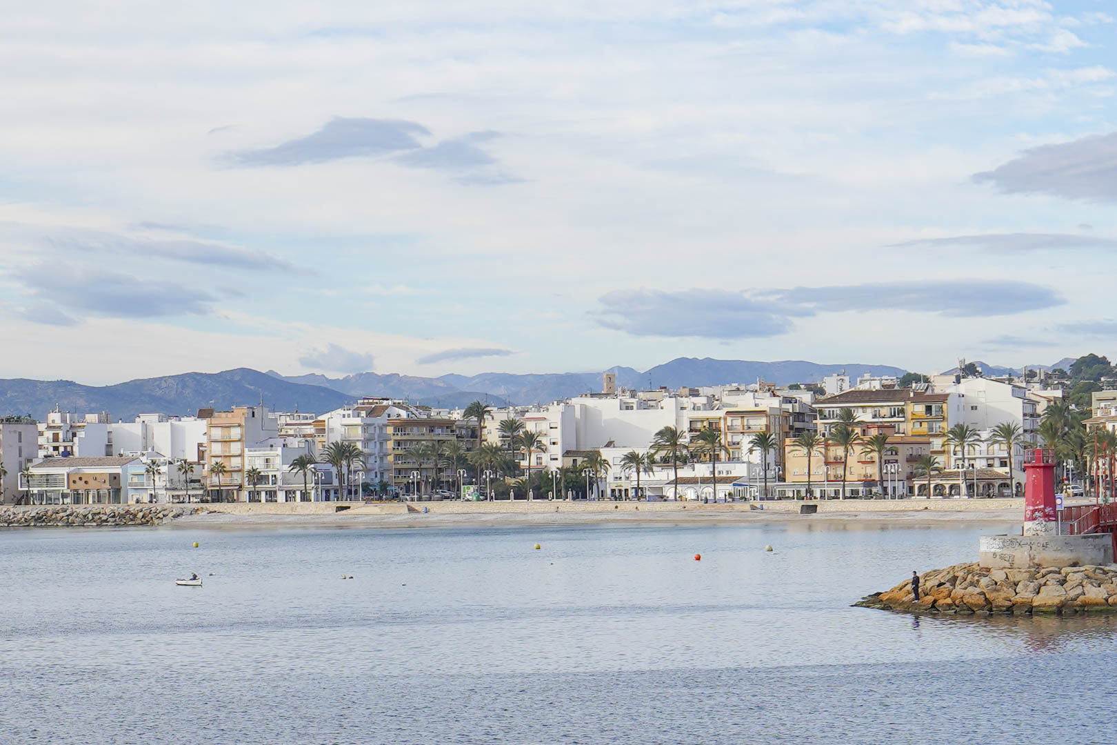 Vista del puerto de Jávea con el faro rojo en primer plano, el paseo marítimo bordeado de palmeras y las montañas de la Marina Alta al fondo.