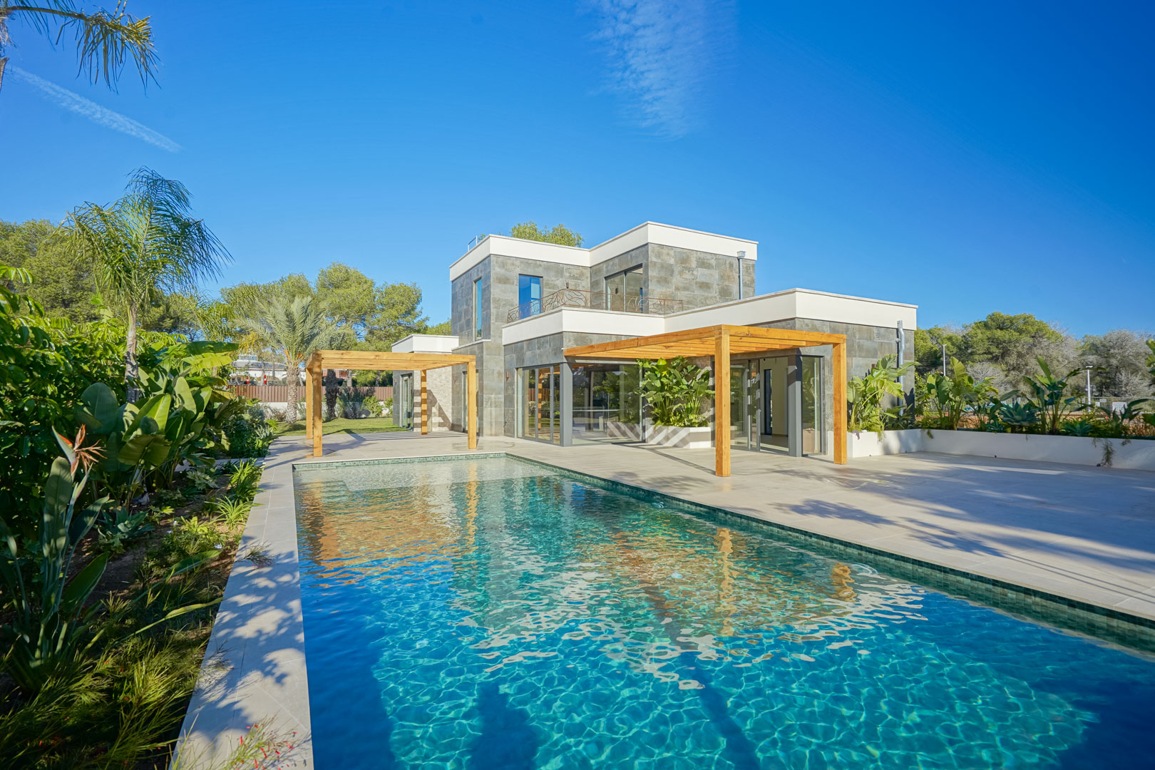 Vista exterior de la villa con piscina de estilo Bali, amplias terrazas y pérgolas de madera de iroko integradas en un jardín mediterráneo.