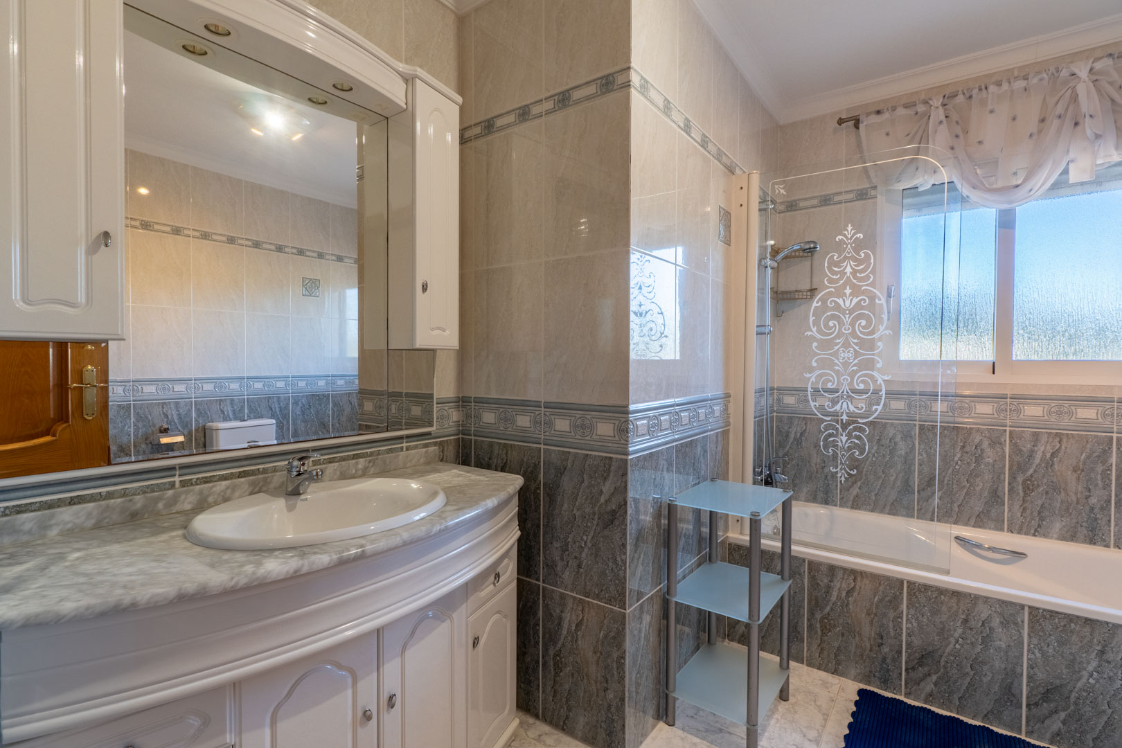Baño completo con encimera de mármol, mueble bajo blanco y bañera con mampara serigrafiada. Paredes revestidas en gres con cenefas decorativas y ventana exterior.
