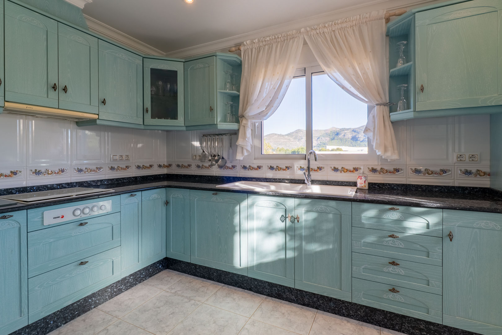 Cocina independiente con mobiliario en tono turquesa, encimera de granito oscuro y ventana con vistas despejadas a las montañas del valle de Jalón.