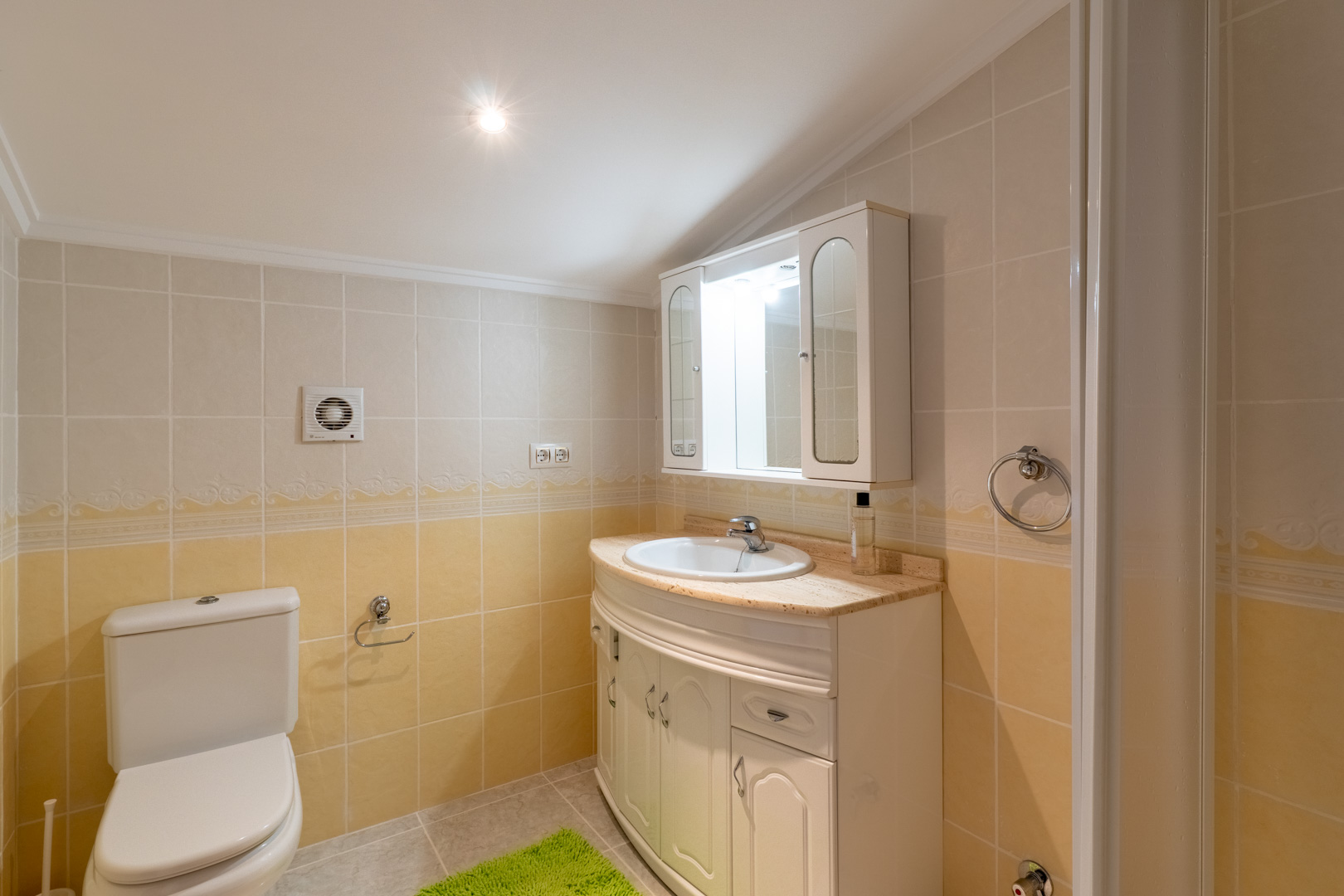 Cuarto de baño con azulejos en tonos crema y amarillo, mueble de lavabo curvo con encimera de mármol y armario superior con iluminación integrada.