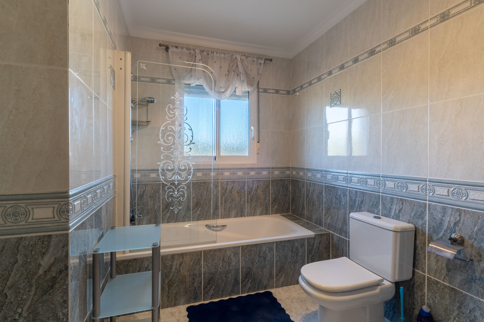 Cuarto de baño con bañera encastrada, mampara de cristal decorada y revestimiento cerámico bitonal con cenefa. Incluye ventana exterior para ventilación natural.