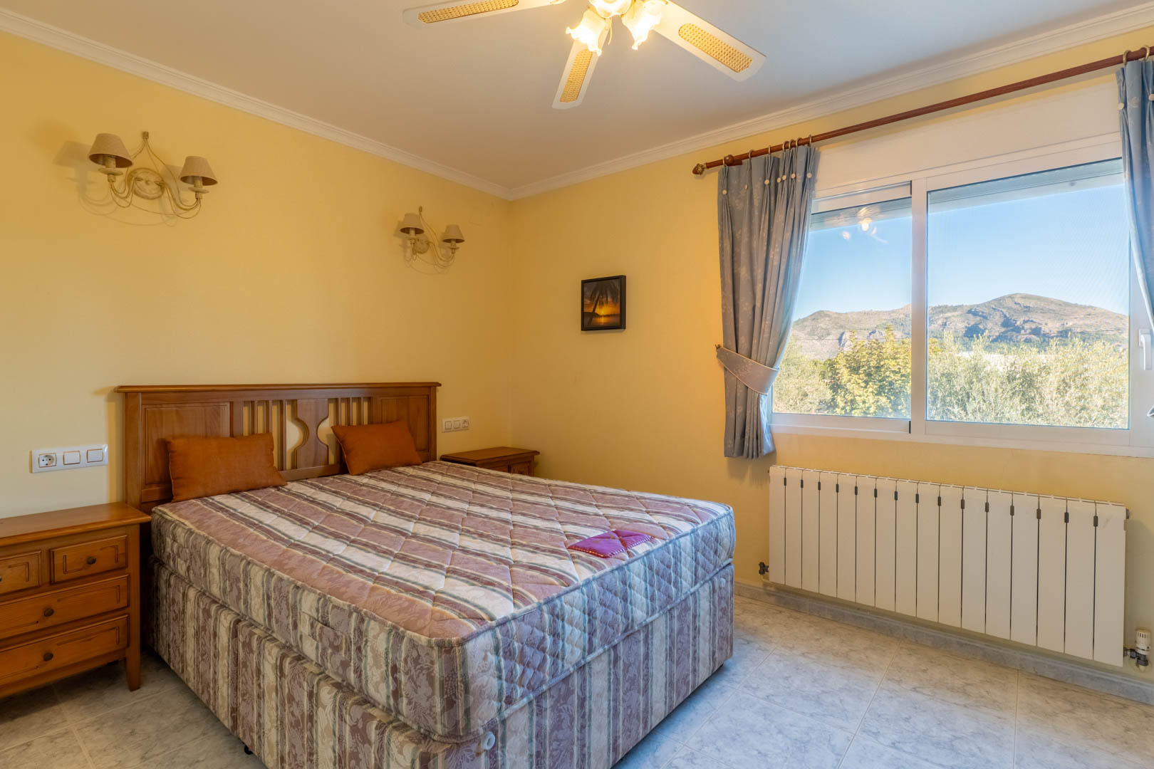 Dormitorio luminoso con mobiliario de madera clásica, ventilador de techo y ventana con vistas despejadas a las montañas y el entorno natural de Jalón.
