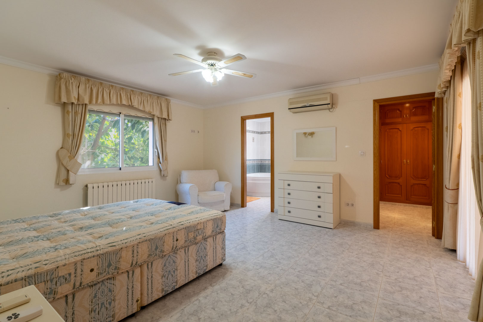 Dormitorio principal con baño en suite y vestidor. Incluye suelos cerámicos, aire acondicionado, ventilador de techo y acceso directo a través de puertas de madera.
