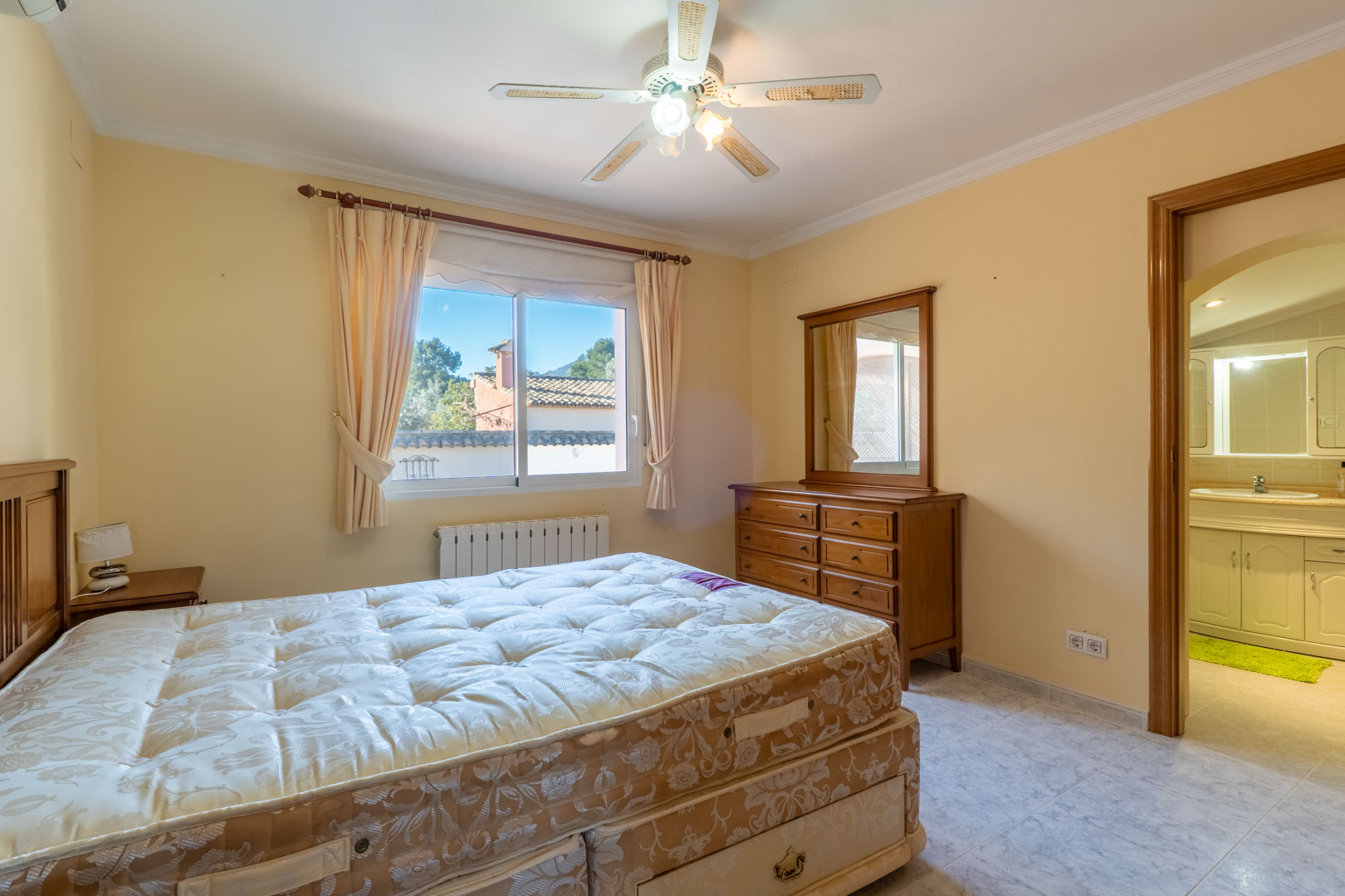 Dormitorio principal con mobiliario de madera, ventilador de techo y acceso directo a un baño en suite. La estancia cuenta con radiador y ventana al exterior.