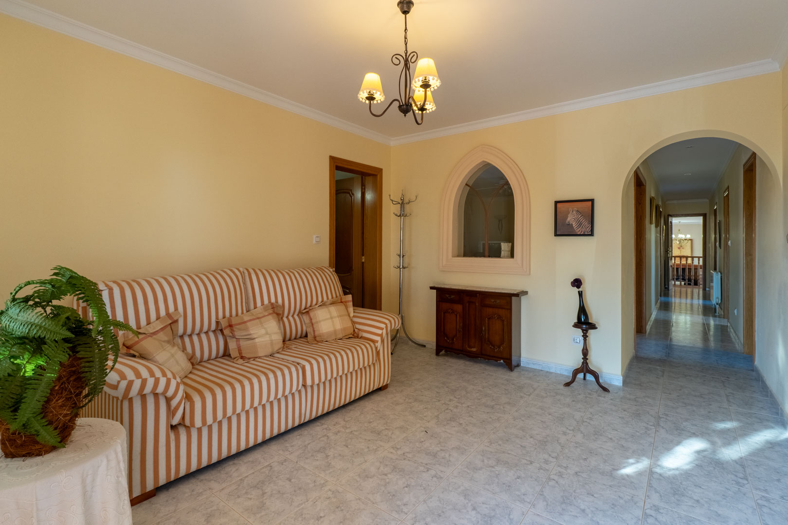 Hall distribuidor con suelos cerámicos, sofá de rayas y un arco que conecta con el pasillo. Destaca una ventana interior decorativa de estilo ojival.