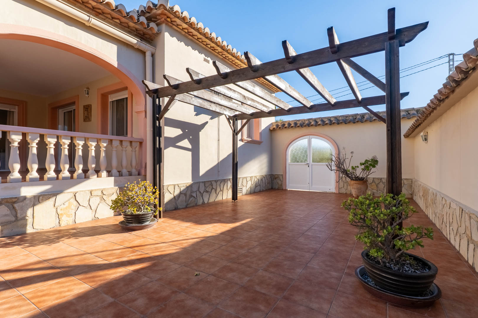 Patio exterior con suelo cerámico, pérgola de madera y muros con zócalo de piedra natural. Incluye acceso a la naya elevada con balaustrada tradicional.