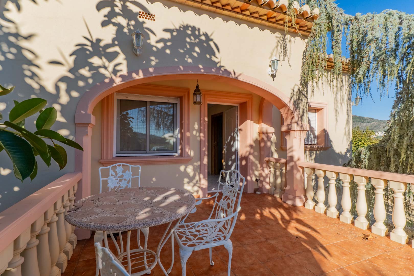 Terraza exterior con arcos de estilo mediterráneo, suelos de terracota y mobiliario de forja blanca, bajo un juego de luces y sombras naturales.