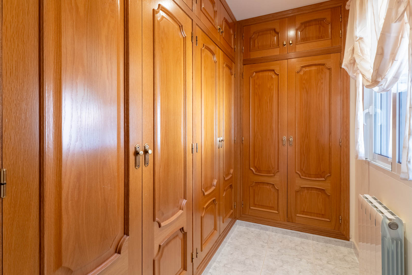 Vestidor con amplios armarios empotrados de madera tallada, suelos de gres cerámico y radiador, iluminado por luz natural lateral.