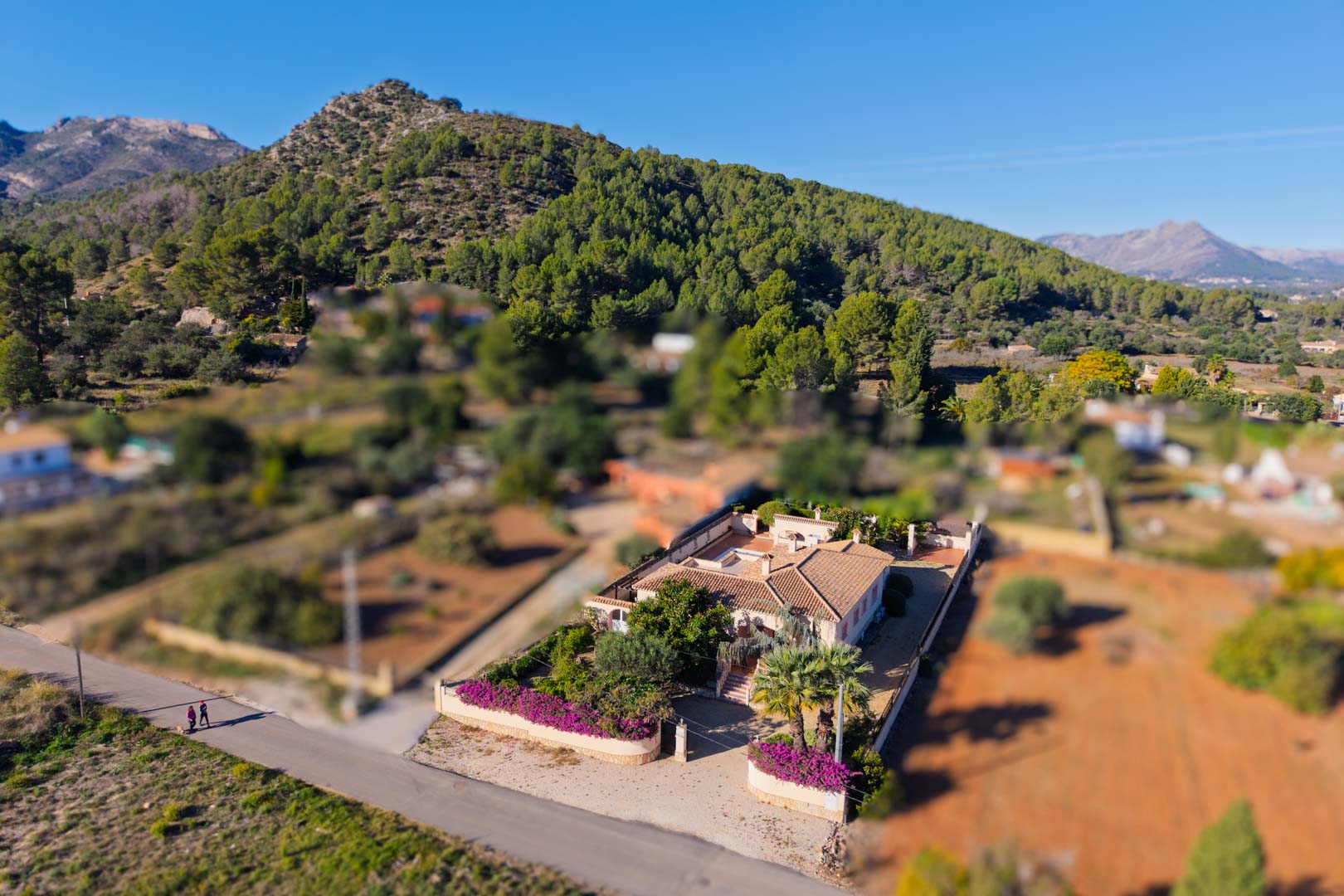 Vista aérea de la villa mediterránea en parcela llana, con tejado de teja tradicional y jardines de buganvillas bajo la falda de la montaña.