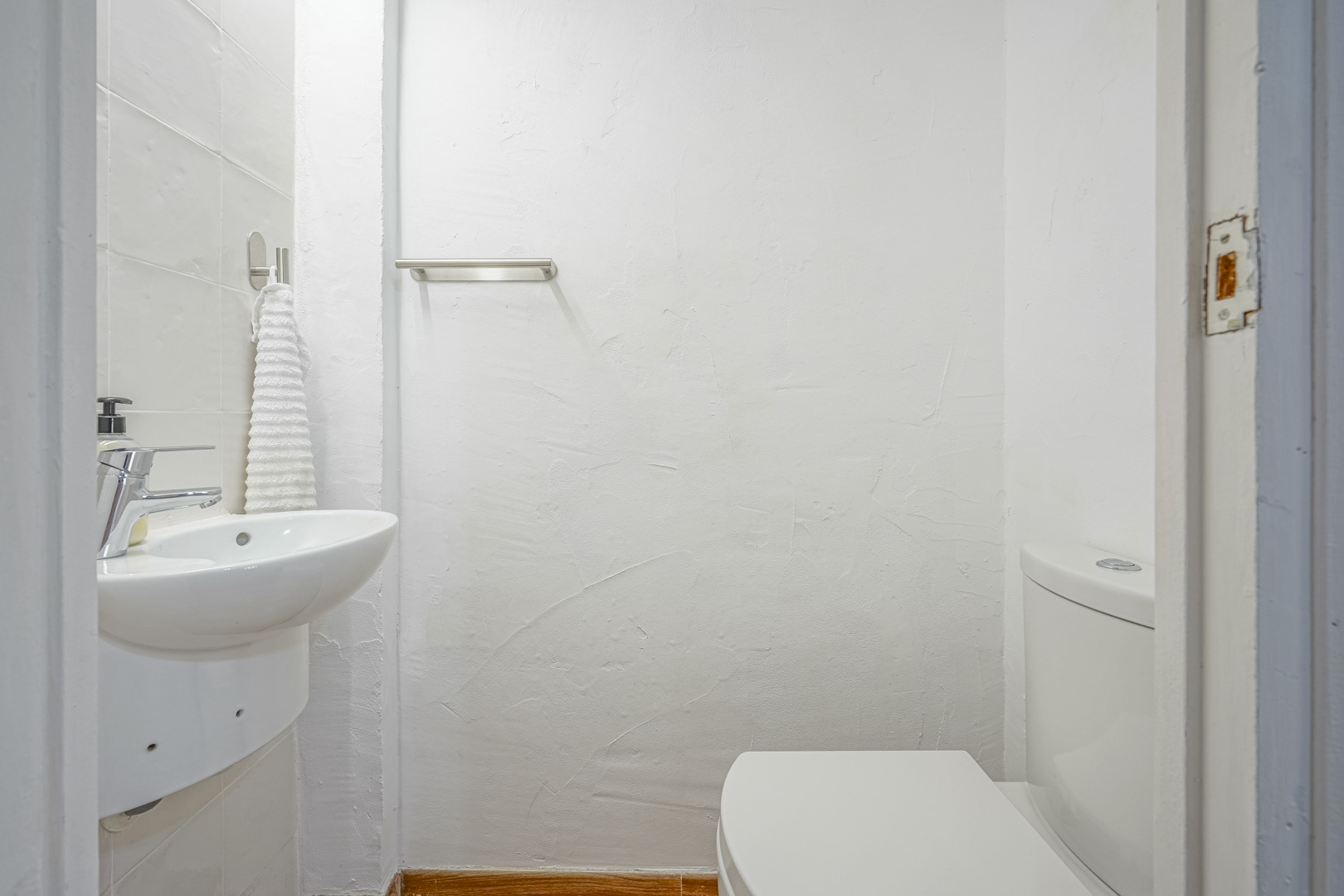 Aseo compacto con paredes de acabado rugoso blanco, lavabo cerámico de pared y grifería cromada. Incluye un toallero metálico y suelos con remate de madera.